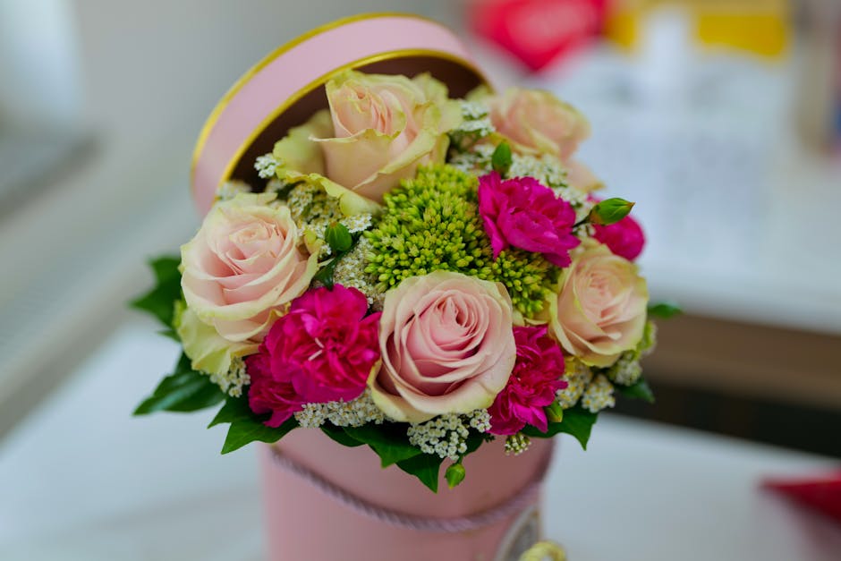 A round floral arrangement inside a light pink cylindrical gift box with a matching pink ribbon, featuring soft pink roses, vibrant fuchsia carnations, green button chrysanthemums, and delicate white 