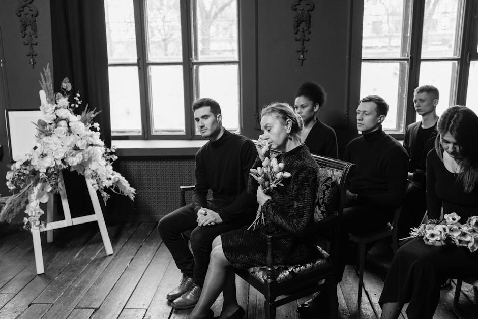 A woman and six young adults sit solemnly in a dimly lit room with large windows, attending a funeral service. The woman, holding a bouquet of tulips, appears emotional, while the others sit quietly w