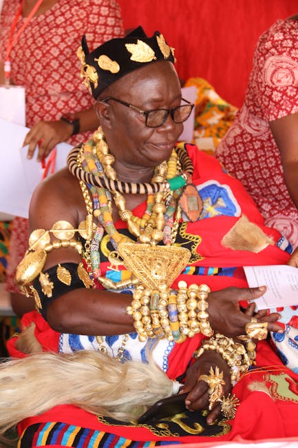 A woman dressed in vibrant traditional clothing decorated with elaborate gold jewelry, including layered necklaces, bracelets, and a headpiece adorned with gold leaves. She is seated indoors, with red