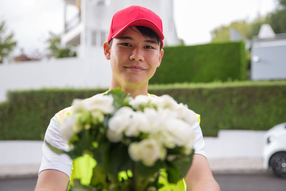 A delivery person wearing a red cap and a high-visibility yellow vest holds out a bouquet of fresh white roses and lilies, with visible green foliage, towards the camera as part of a floral gift deliv