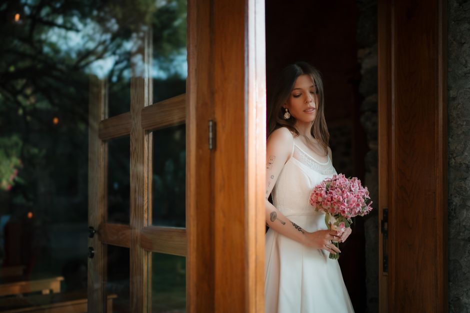 A young woman with shoulder-length brown hair, dressed in a white lace and satin wedding dress, stands inside a rustic stone building, holding a bouquet of pink hydrangeas. She is partially visible th